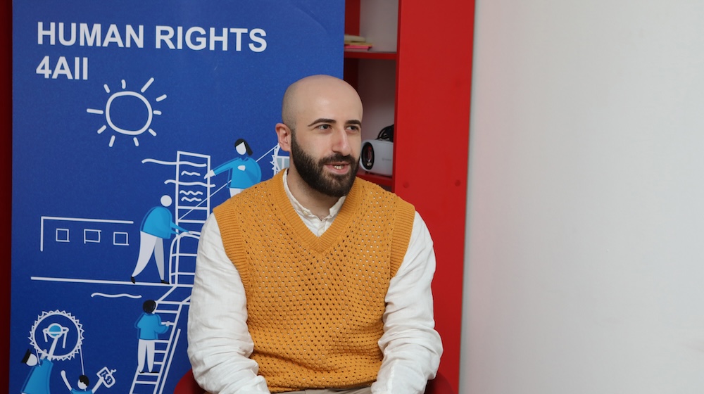 Beka Gabadadze in an orange sweater and white shirt speaking with a blue UNDP banner behind him that reads "Human Rights 4 All"