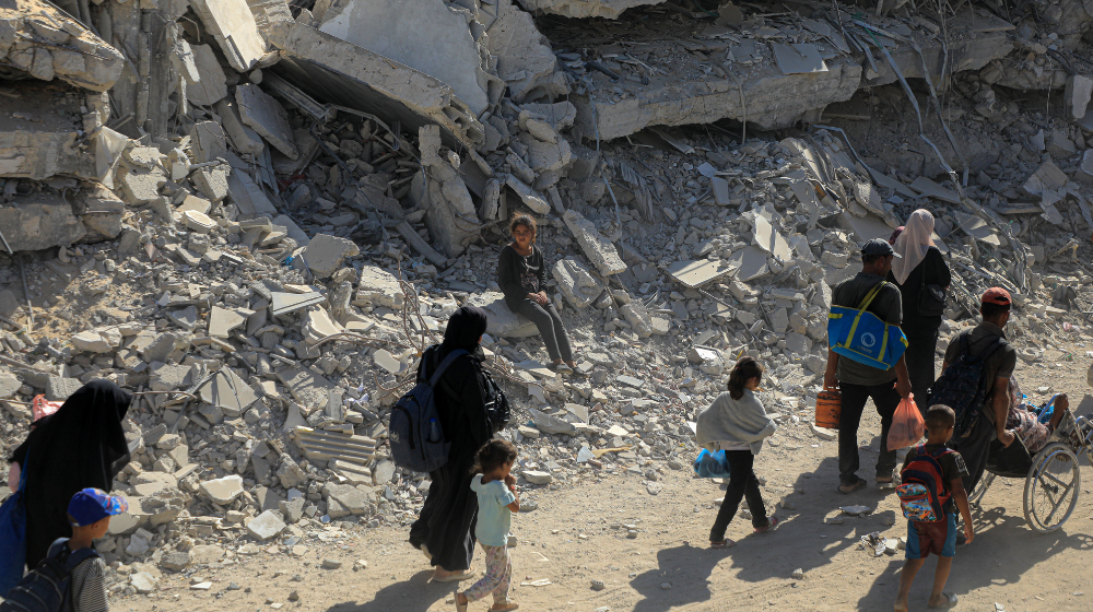  Un groupe de personnes longe des bâtiments en ruine, portant quelques maigres affaires. Une petite fille est assise sur un tas de gravats et les regarde passer