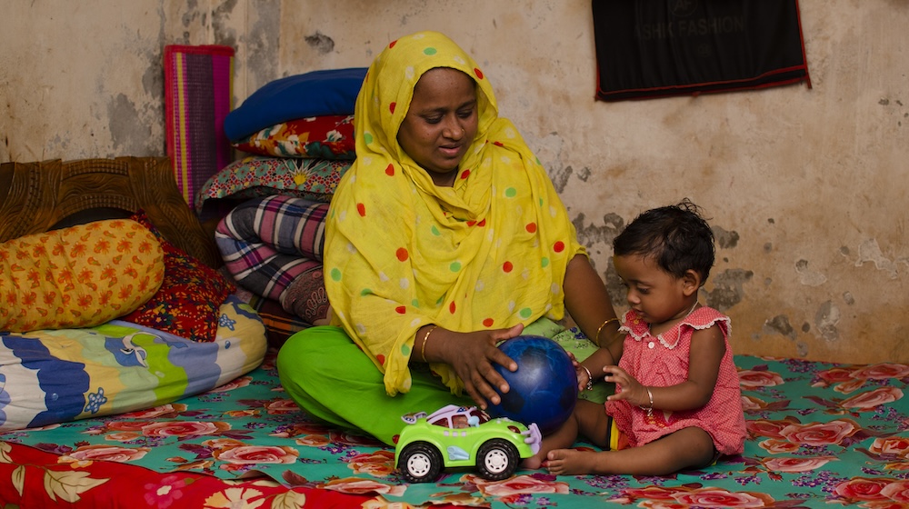 A mother plays with her child while sitting on a bed.