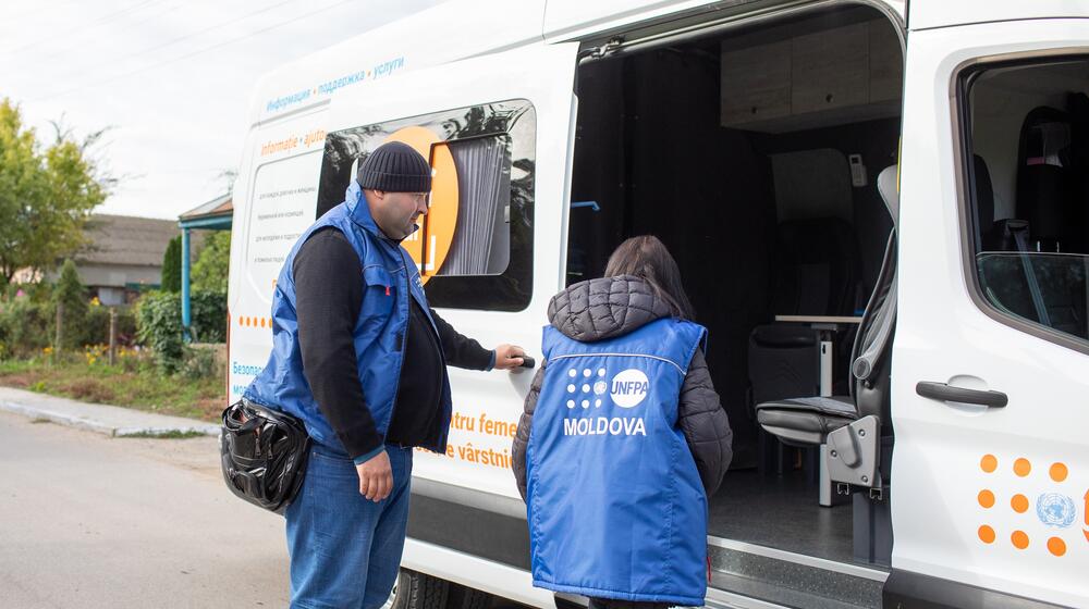 Dos trabajadores del UNFPA junto al vehículo de un gran espacio seguro móvil que consta de asientos y una mesa.