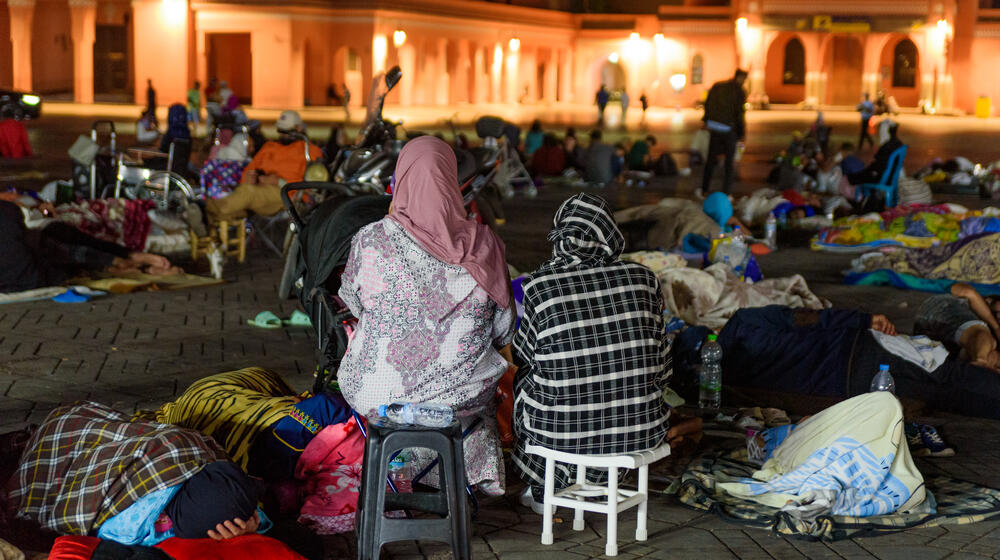 Deux personnes sont assises sur des petites chaises. Elles sont dehors, en compagnie de nombreux autres individus qui dorment à même le sol.