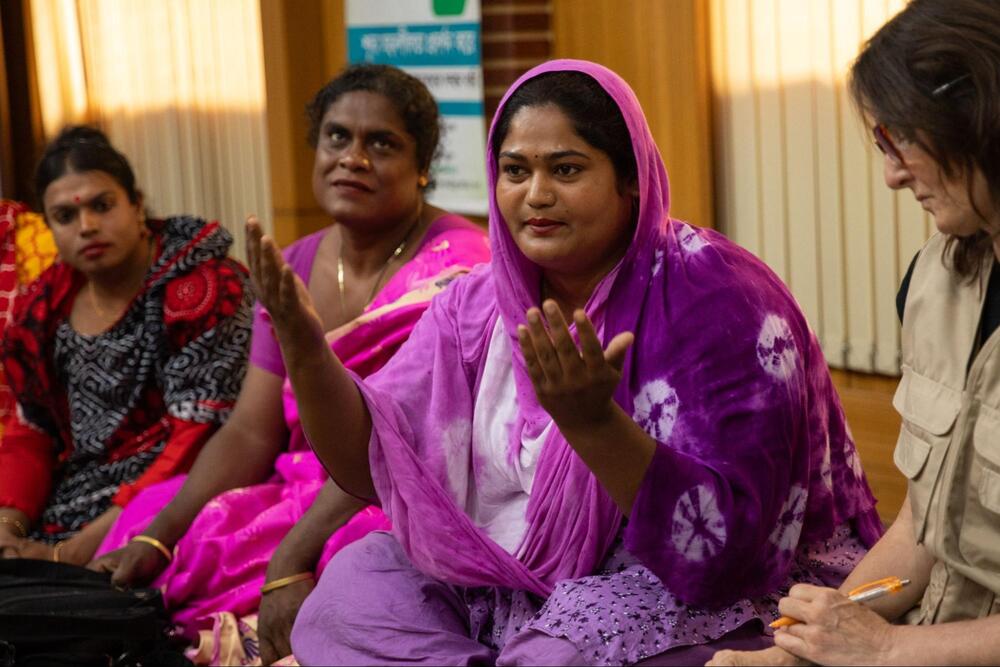 Maimouni, miembro del grupo hijra de Bangladesh, vestido con ropa rosa y púrpura, sentado, habla entre un grupo.