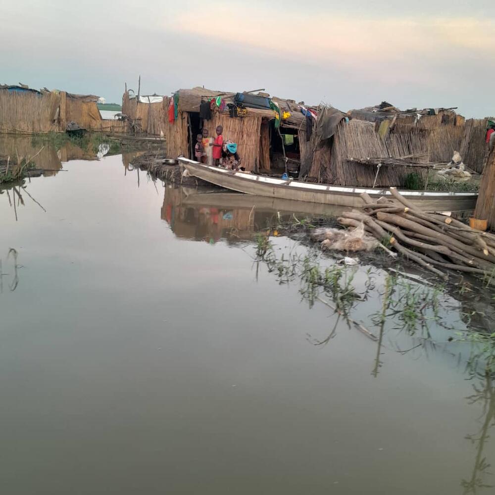 A family stands in the doorway of a thatch home. They are reflected in the water surrounding their home.