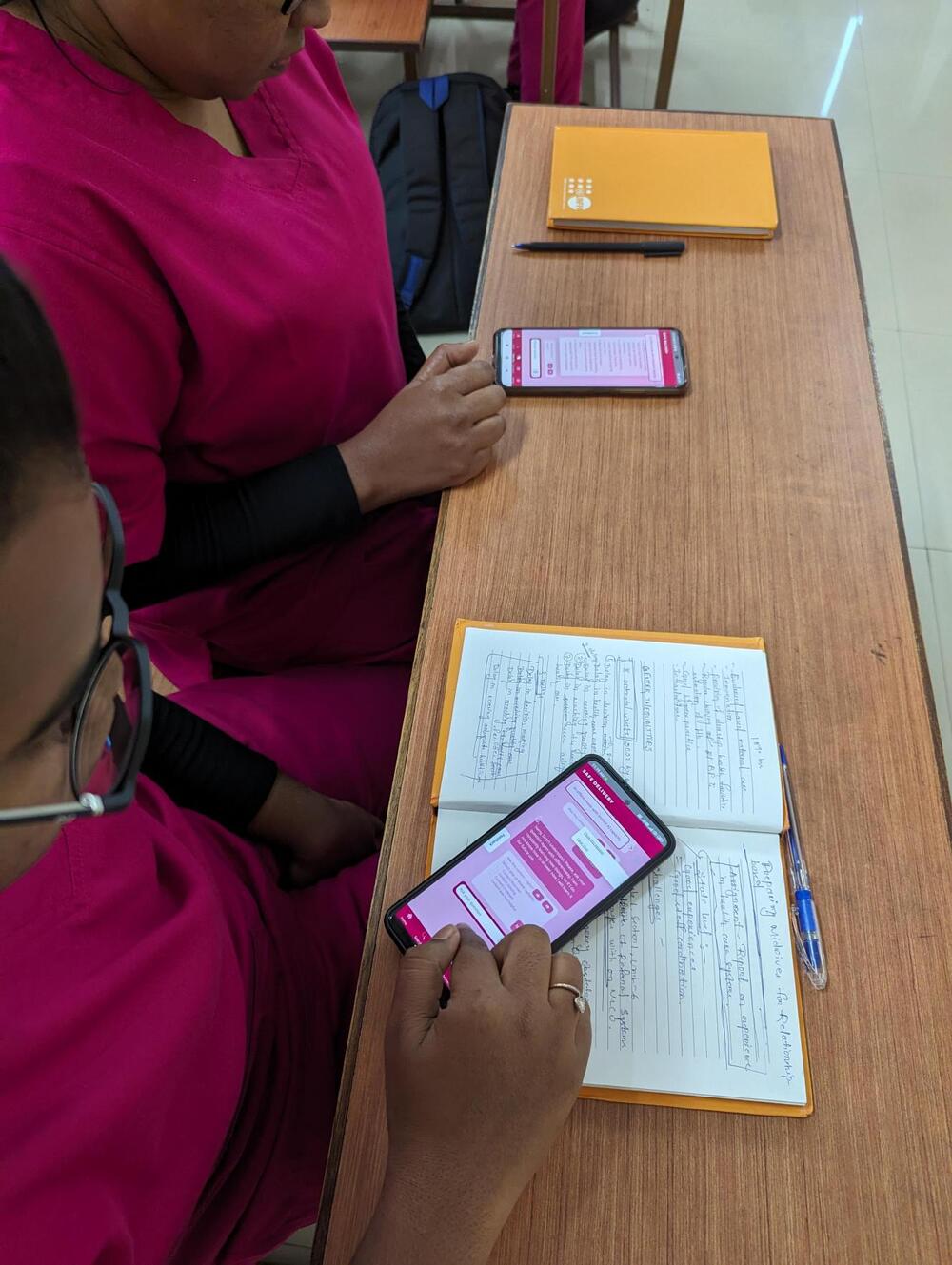 Two women sit at a desk and scroll through their phones with notebooks beside them.