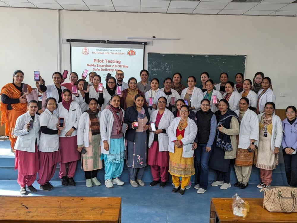 A large group of nurses and midwives stand in a classroom and hold up their phones to the camera.