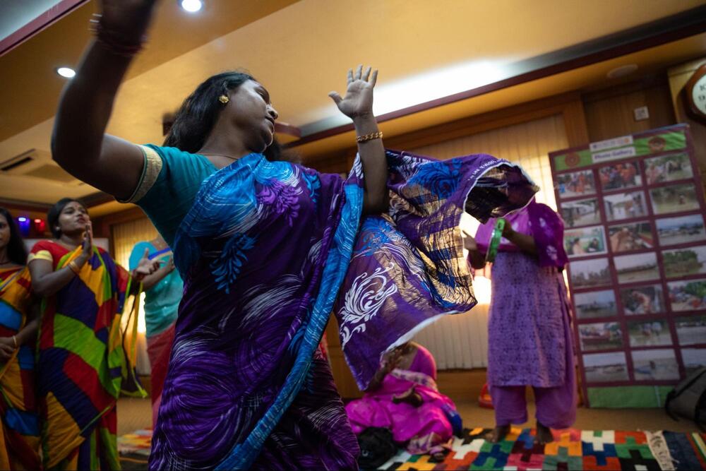 Maimouni, membre de la communauté hijra du Bangladesh, vêtue de rose et de violet, assise en train de discuter avec un groupe de personnes.