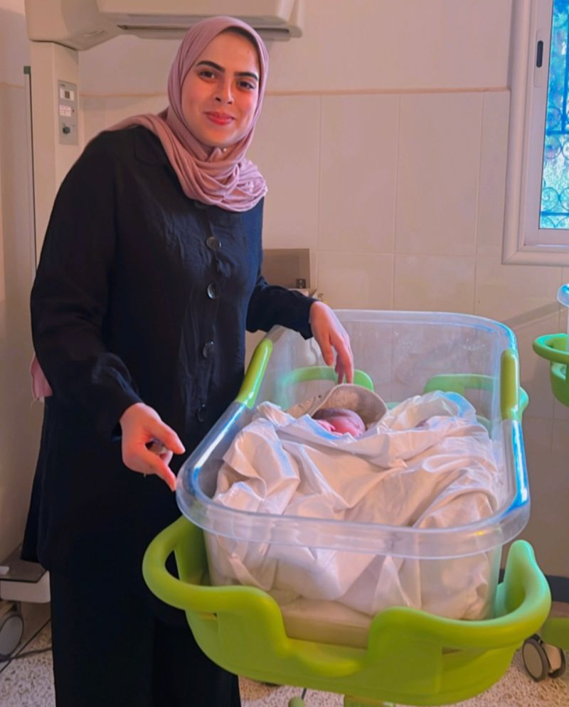 A midwife smiles as she stands next to a newborn baby in a hospital.