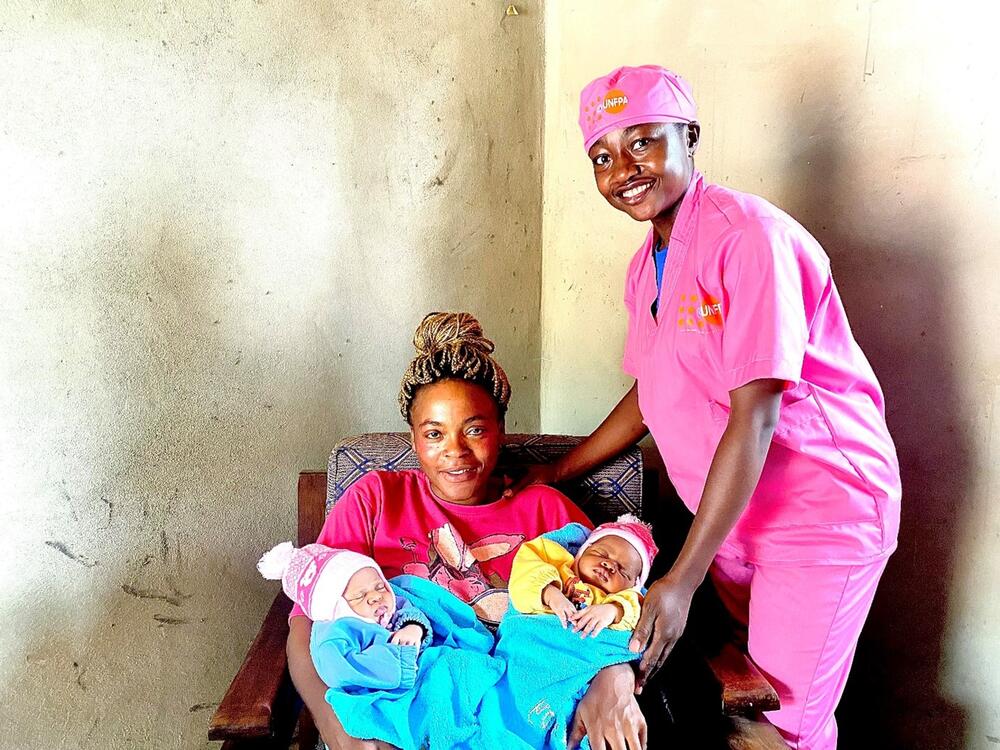 A midwife in a pink uniform smiles while standing next to a seated mother holding two newborn babies.