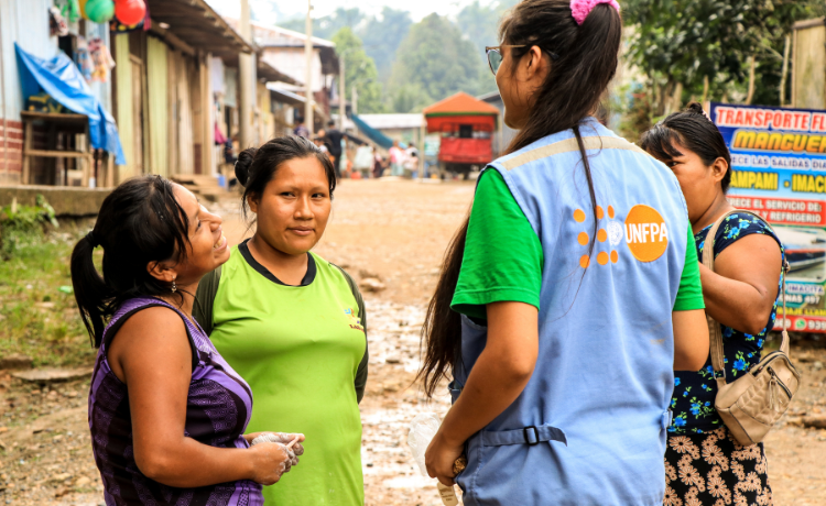 Cuatro mujeres charlan en un círculo. Una lleva un chaleco azul con el logotipo del UNFPA. Todas están de pie en una calle del pueblo.