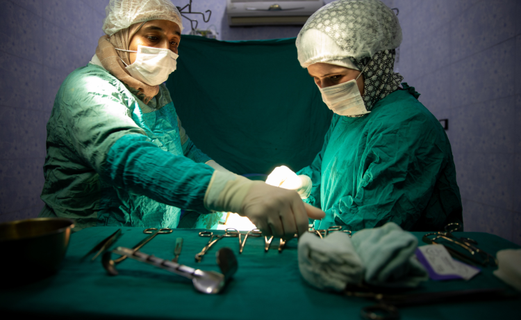 Deux agentes de santé en blouses chirurgicales vertes, portant masques et gants, se tiennent près d’une table où sont disposés des instruments