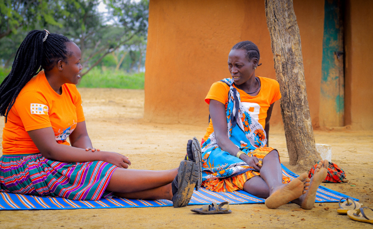  Une femme portant un t-shirt orange floqué du logo de l’UNFPA parle à Mme Telo, qui porte également un haut orange. Elles sont assises sur une couverture à rayures dans une cour.