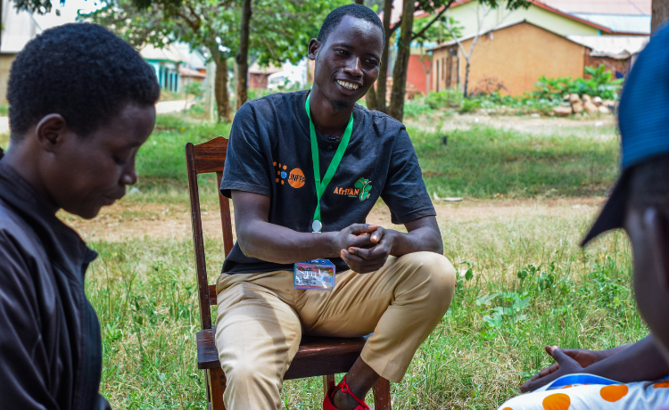  A man in a black t-shirt with an orange UNFPA logo sits on a chair in the shade of a courtyard. He is speaking to other young people.