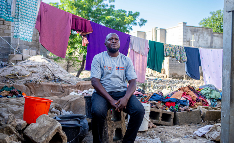 A man sits on a cinderblock in front of rubble from a flood and fabric hanging from a clothing line. He wears a gray t-shirt with the UNFPA logo.