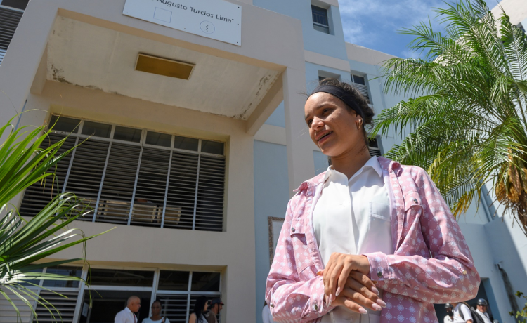 A young woman wearing a white shirt and pink jacket stands outside a health clinic in Cuba.