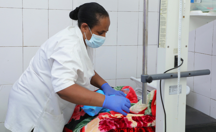 Una mujer con bata blanca y mascarilla y guantes azules atiende a un recién nacido envuelto en mantas de colores en una sala del hospital