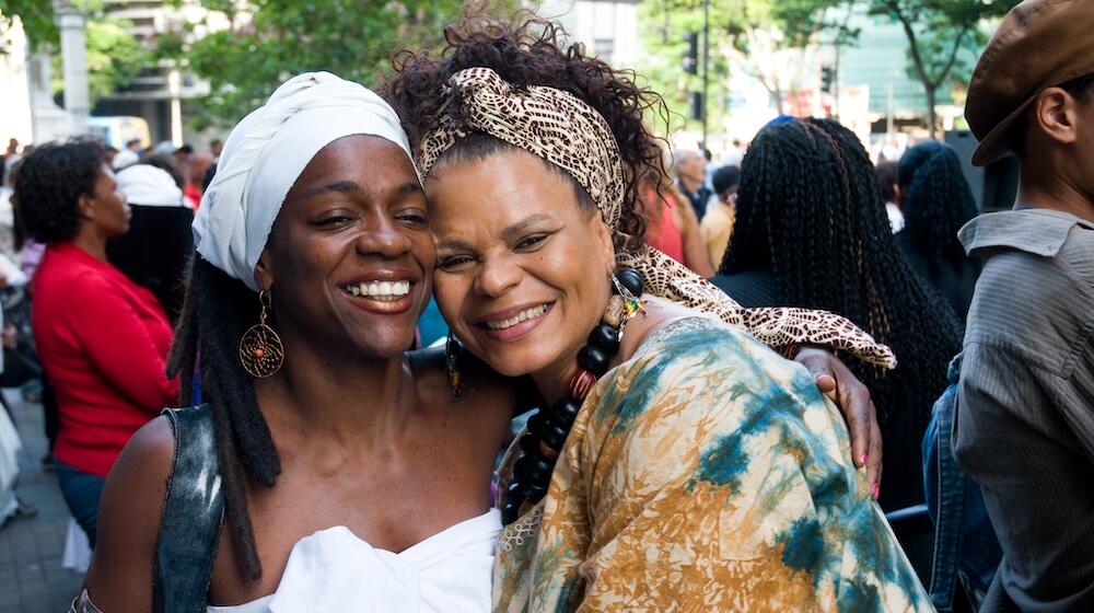 Two women of African descent embrace and look towards the camera. 