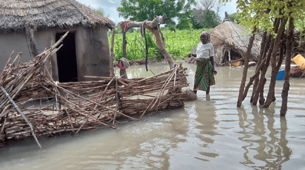 Une femme se tient debout dans l’eau d’une crue, près d’une clôture en bois effondrée et d’une maison au toit en chaume