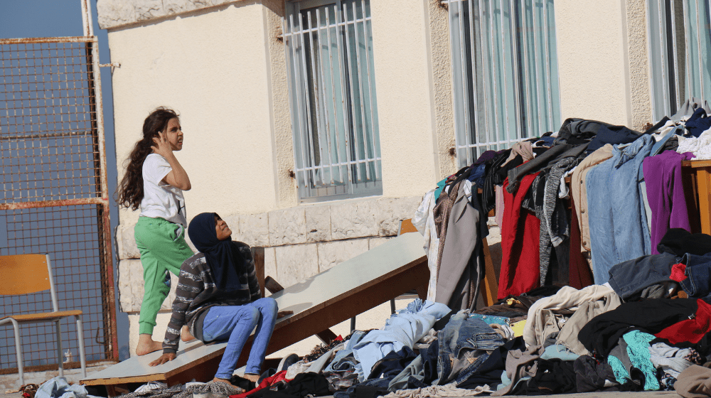 Girls sit outside a public school serving as a shelter. Near them is furniture covered in colorful piles of clothes.