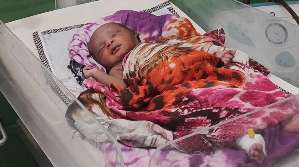  A newborn baby is wrapped in a colourful headscarf in a hospital bassinet.