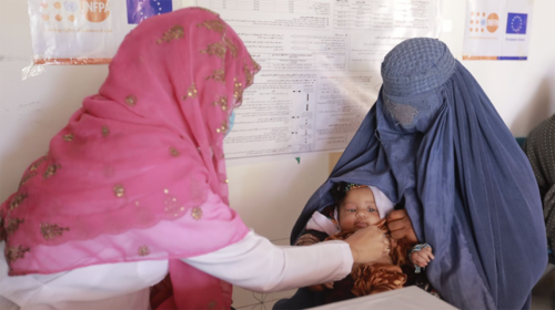 Two women sit opposite each other. The woman in a blue burka holds a small baby. The second woman in a pink headscarf reaches over to touch baby's face affectionately