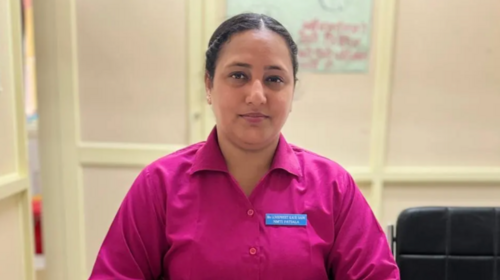 Une femme portant un uniforme rose d’infirmière est assise à un bureau devant un livre et un téléphone.