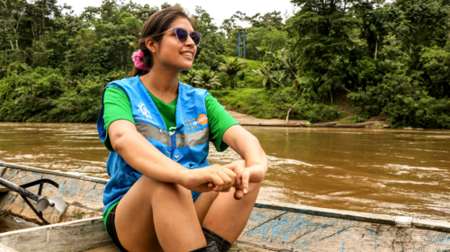 A young woman sits in a wooden rowboat on a river surrounded by jungle.