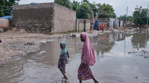  Girls wearing pale blue and pink headscarves walk through a flooded street.